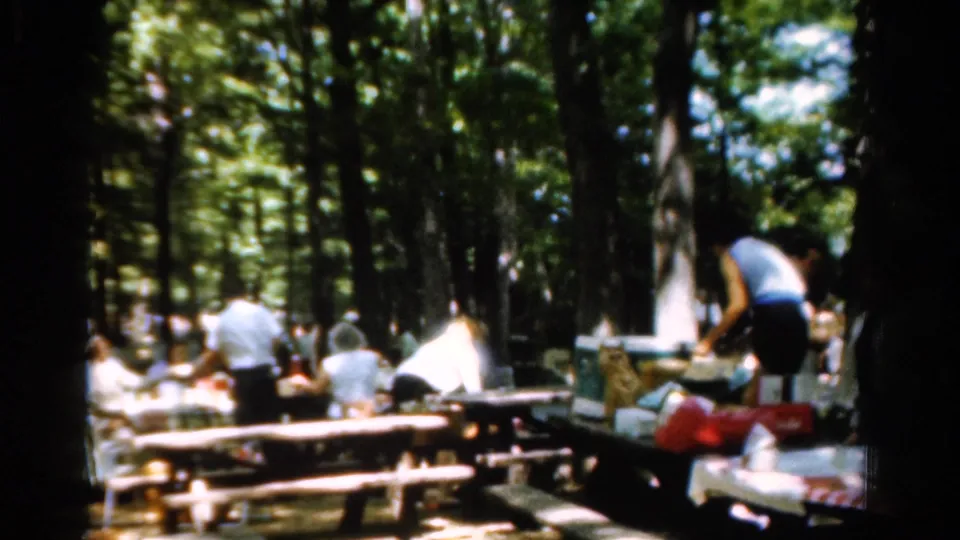 Home-movie footage shows adults at picnic tables in a shaded California park, with supplies spread across benches and tables.