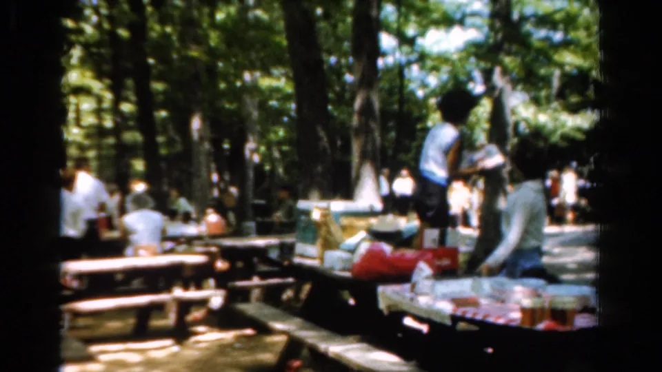 1954 California park picnic scene with adults near wooden tables, shaded trees, red bags, and food set out on a cloth.