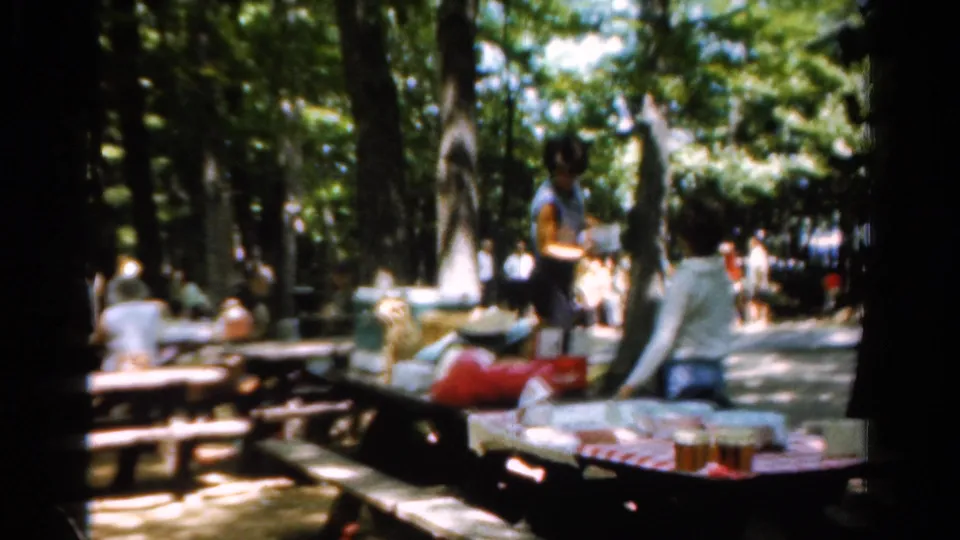 Soft 8mm frame from a 1954 California picnic, showing adults beside a table loaded with bags, food, and drinks.