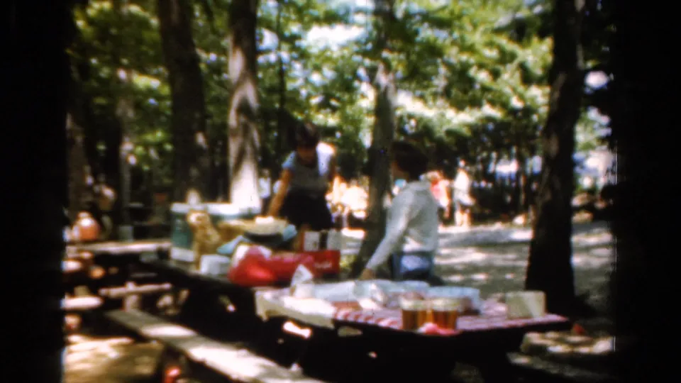 1954 California home-movie view of adults at a shaded picnic table with food, drinks, bags, and trees behind them.
