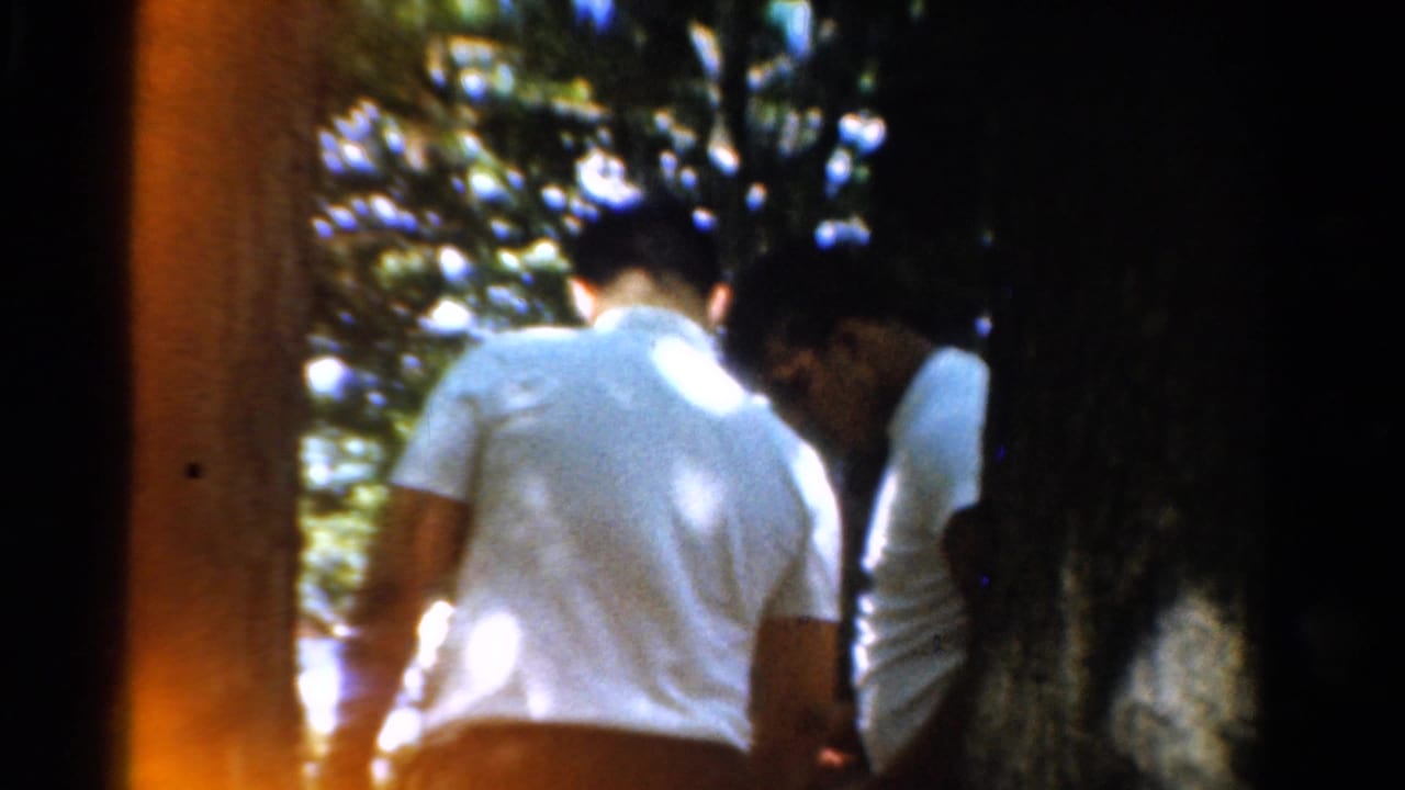 In 1954 California 8mm footage, men in white shirts move through a wooded area with dappled sunlight and tree trunks obscuring them.