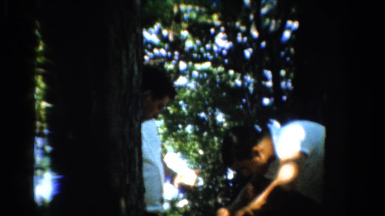 Men in white shirts stand under trees, with one bending toward something near his hands.