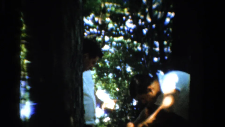 1954 California home-movie frame showing adult men in white shirts gathered in a wooded area, partly obscured by tree trunks and shade.