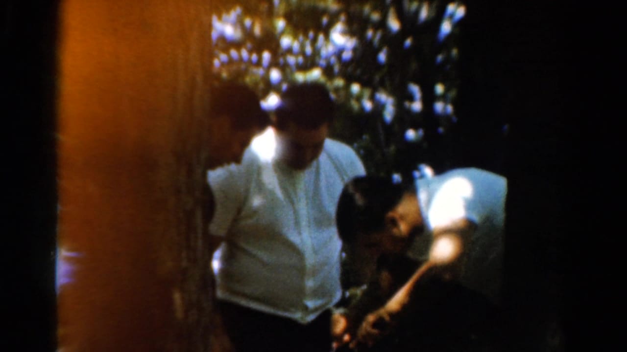 1954 California frame with three adult men in shirts and trousers focused downward in a wooded outdoor setting.