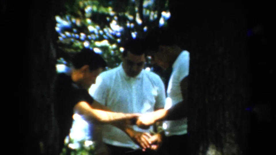 1954 California home-movie frame of adult men under trees, one extending an arm toward a small object or food item being held.