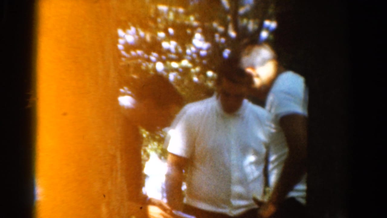 1954 California 8mm frame of adult men in light shirts standing in a wooded area with dappled sun and orange edge flare.