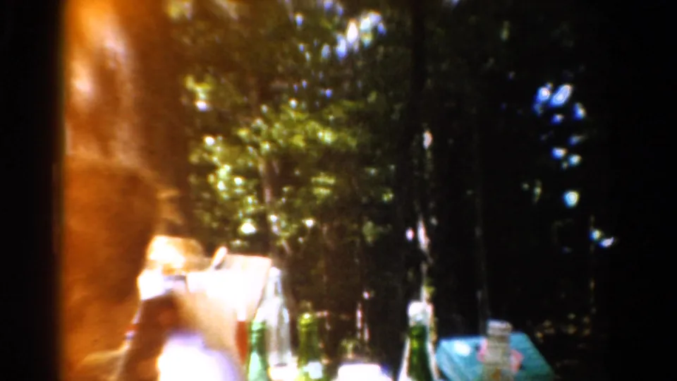 1954 California campsite frame with a person near a table, bottles in the foreground, and trees behind.
