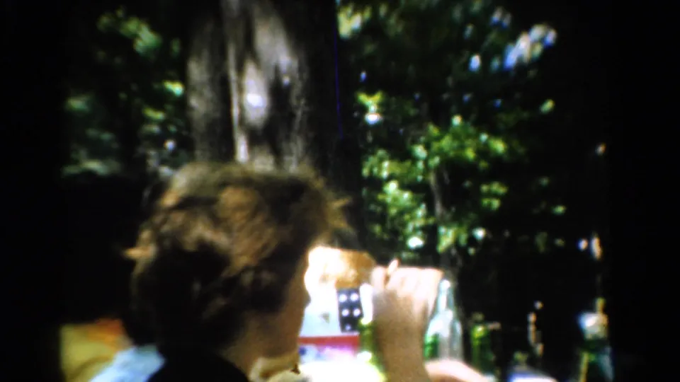 A curly-haired person in profile appears to eat or drink at a shaded California campsite table in 1954 home-movie footage.