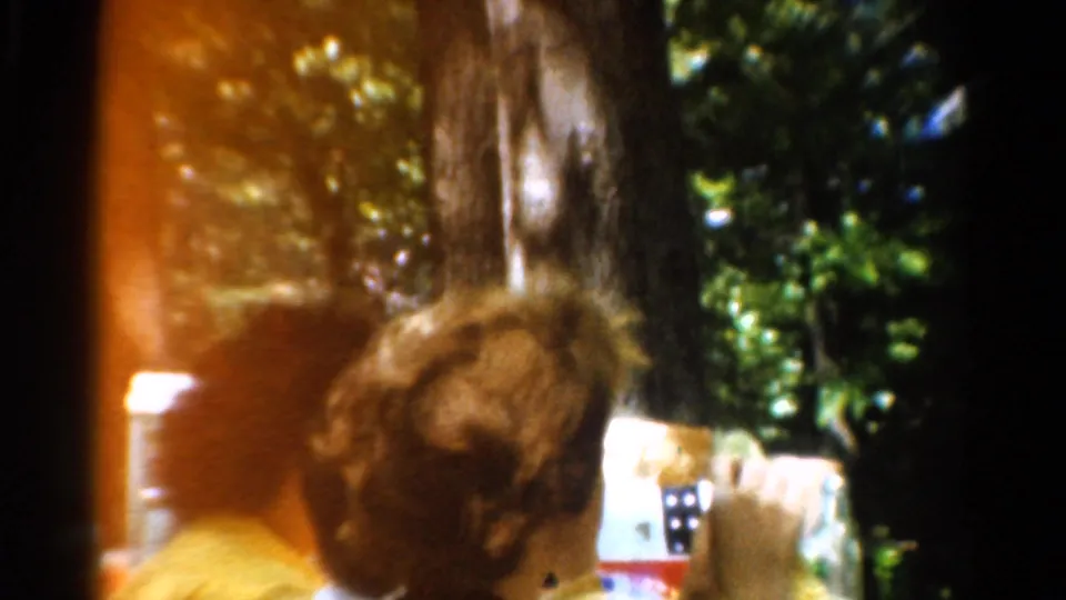 1954 California 8mm frame showing the back of a curly-haired person at an outdoor meal, with bottles and trees softened by film exposure.