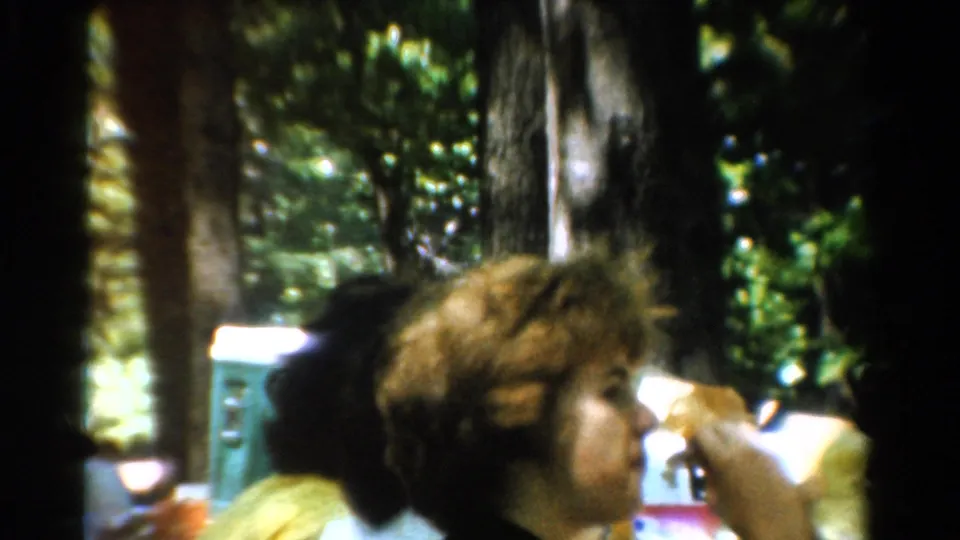 A curly-haired person and another partially visible person sit at a shaded outdoor table in 1954 California camping footage.