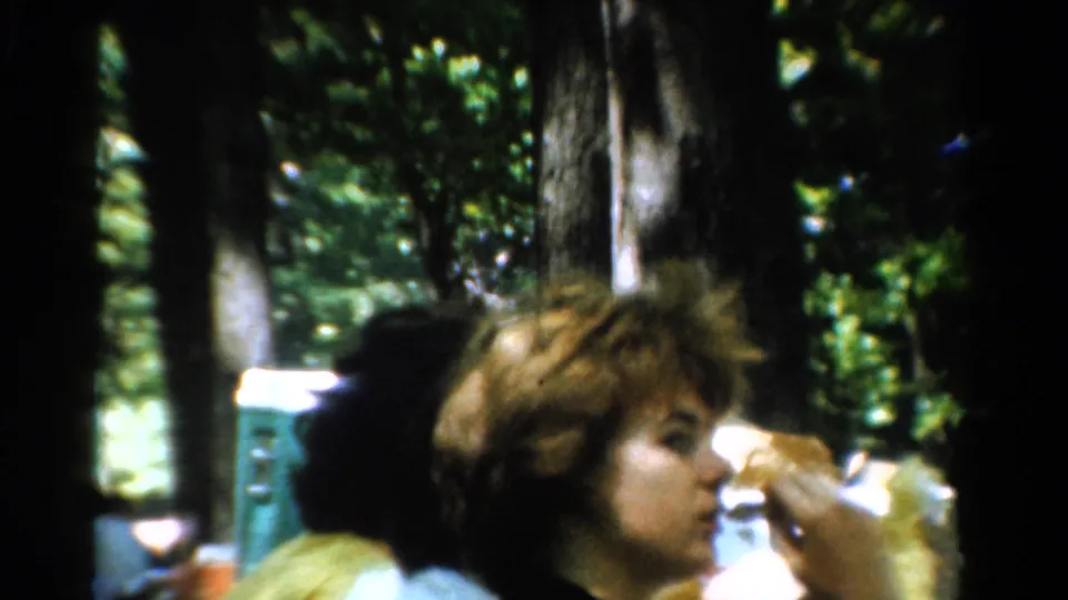 1954 California 8mm frame of adults at a wooded picnic table, with one person eating a sandwich in close foreground.