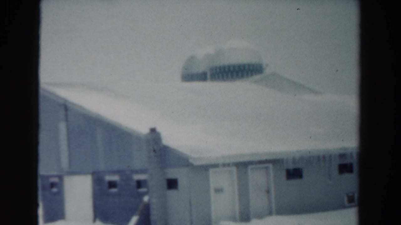 1954 Toledo winter frame with a low snow-covered building, hanging icicles, small windows, and a distant domed or rounded roof form.