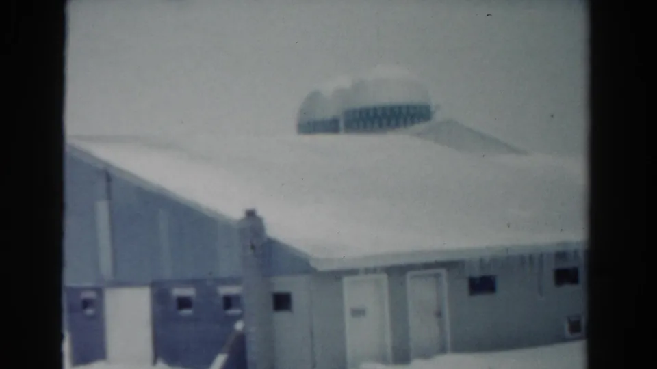 1954 Toledo winter frame with a low snow-covered building, hanging icicles, small windows, and a distant domed or rounded roof form.