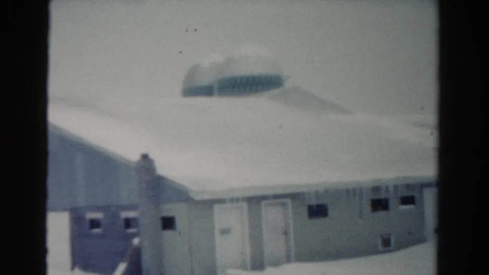 1954 Toledo 8mm frame showing a closer view of the snow-loaded roof, icicles, pale wall, doors, and dark window openings.