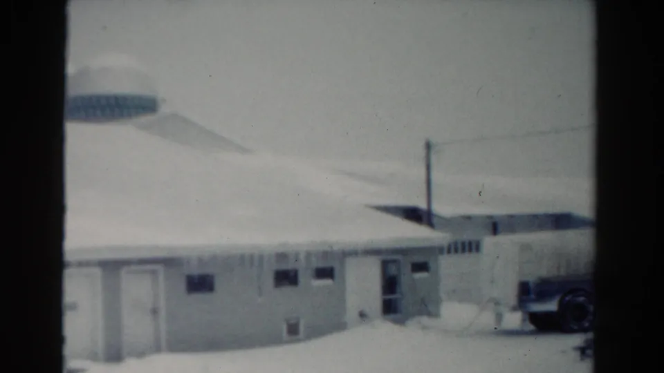1954 Toledo 8mm frame showing the snowy building, icicles, utility pole, rounded background structure, and part of a parked vehicle at right.