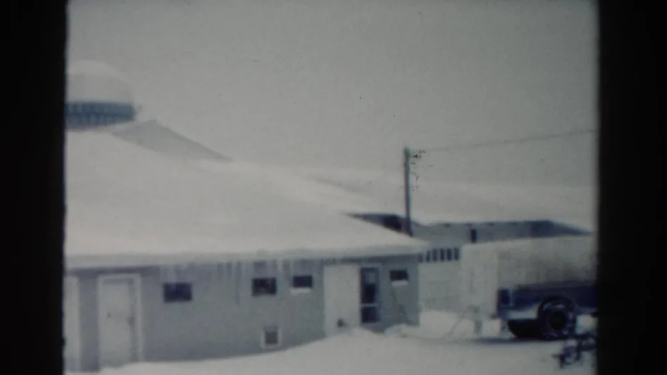 1954 Toledo home-movie frame showing a snow-covered low building, power pole, curved roofline, and part of a parked dairy truck.
