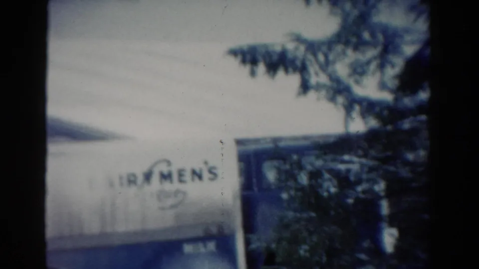 1954 Toledo home-movie frame with a white Fairmen's Dairy truck and partial MILK marking behind evergreen trees.