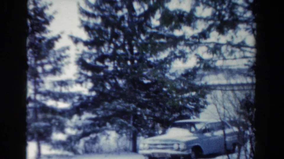 1954 Toledo home-movie frame showing a light blue station wagon parked in snow behind evergreen trees.