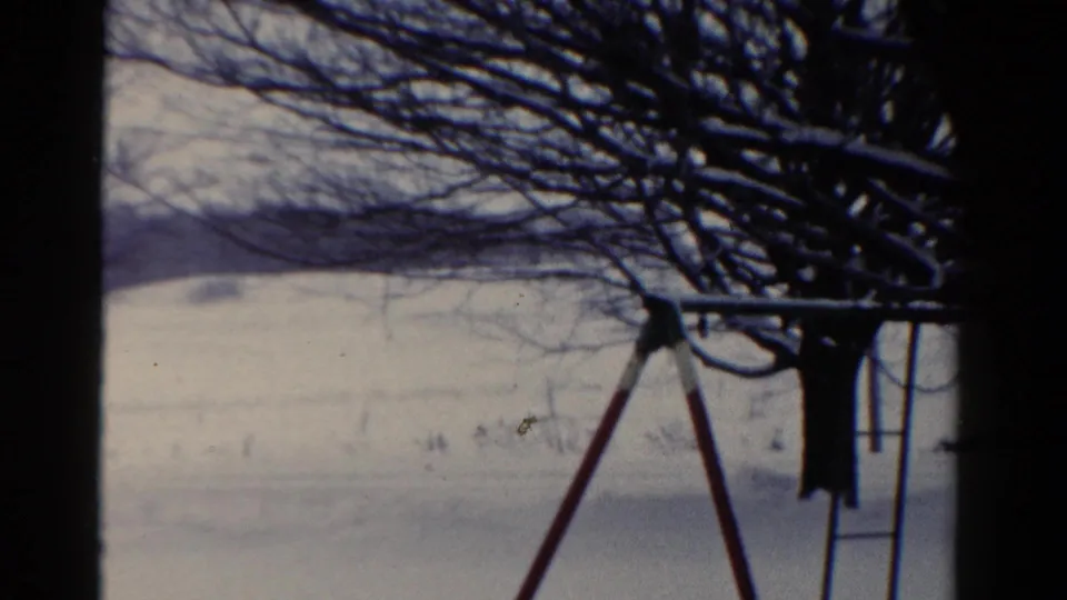 1954 Toledo winter frame showing a snow-covered playground area, bare branches, and metal play equipment at the right edge.