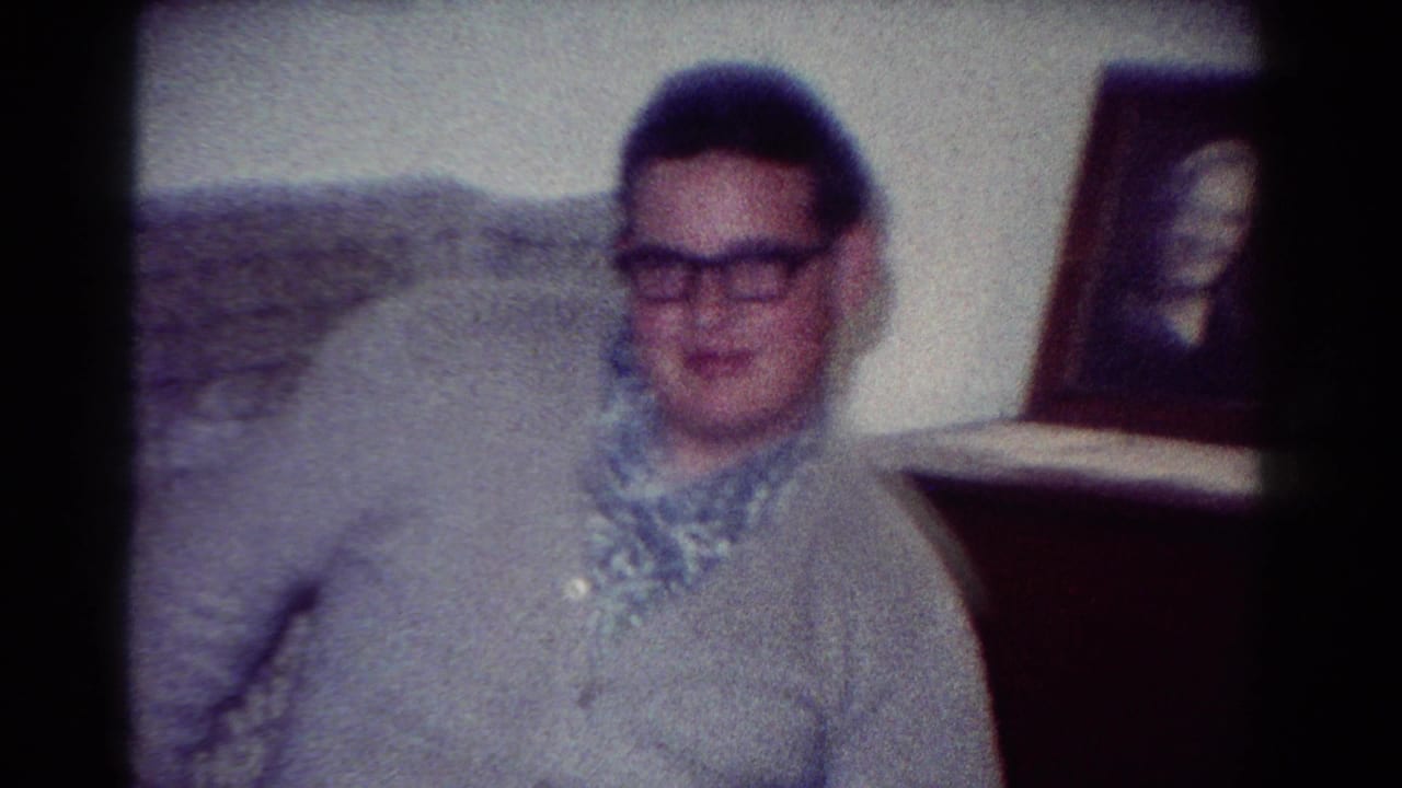 In the supplied 1954 Toledo home-movie context, a young man in glasses sits awkwardly near a framed portrait in a private interior.