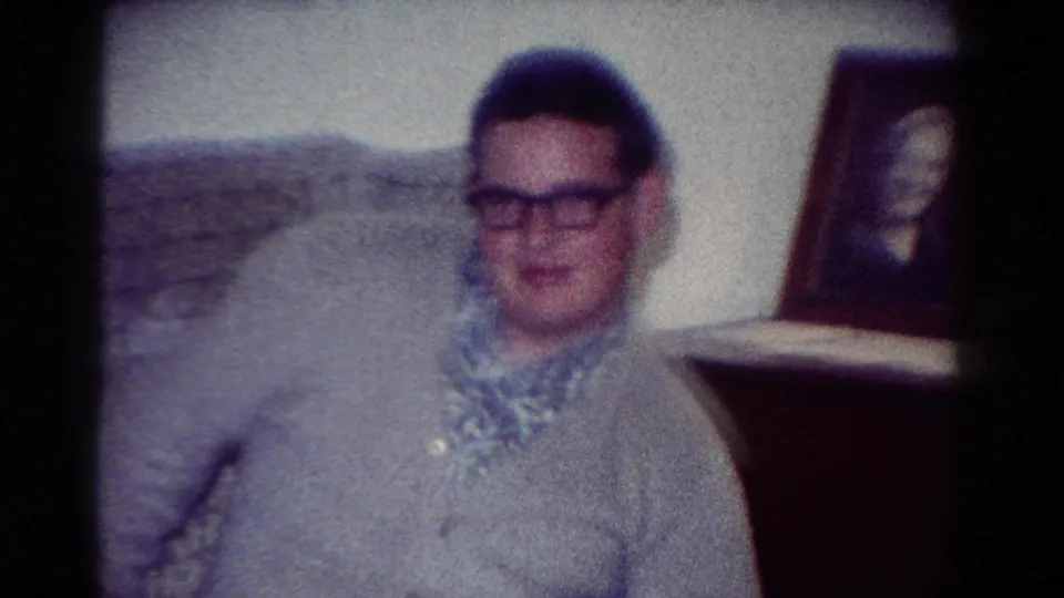 In the supplied 1954 Toledo home-movie context, a young man in glasses sits awkwardly near a framed portrait in a private interior.