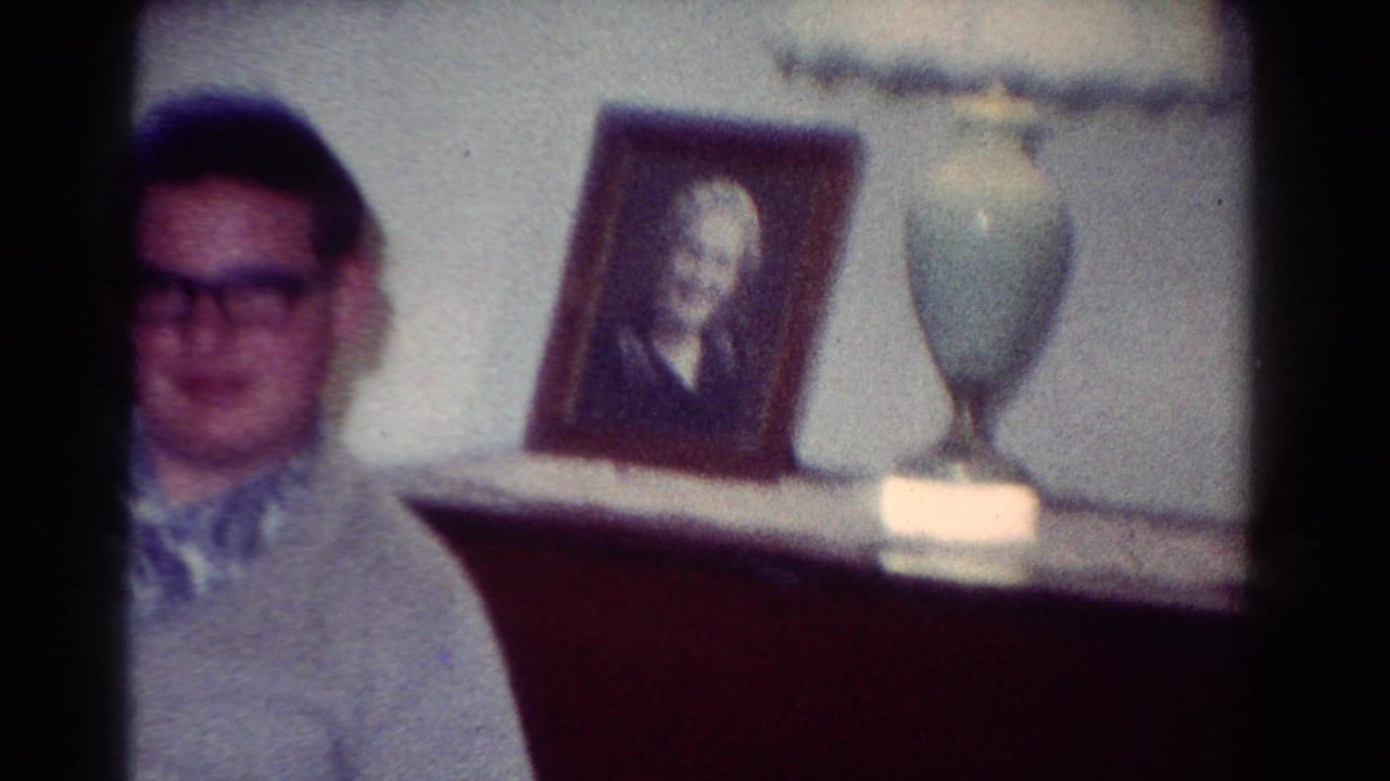 A young man in glasses is partly framed by dark furniture, with a portrait and pale lamp visible in the 1954 Toledo interior.