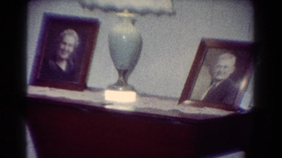 Two framed older-adult portraits and a pale blue lamp sit on a dark cabinet in the supplied 1954 Toledo home-movie setting.