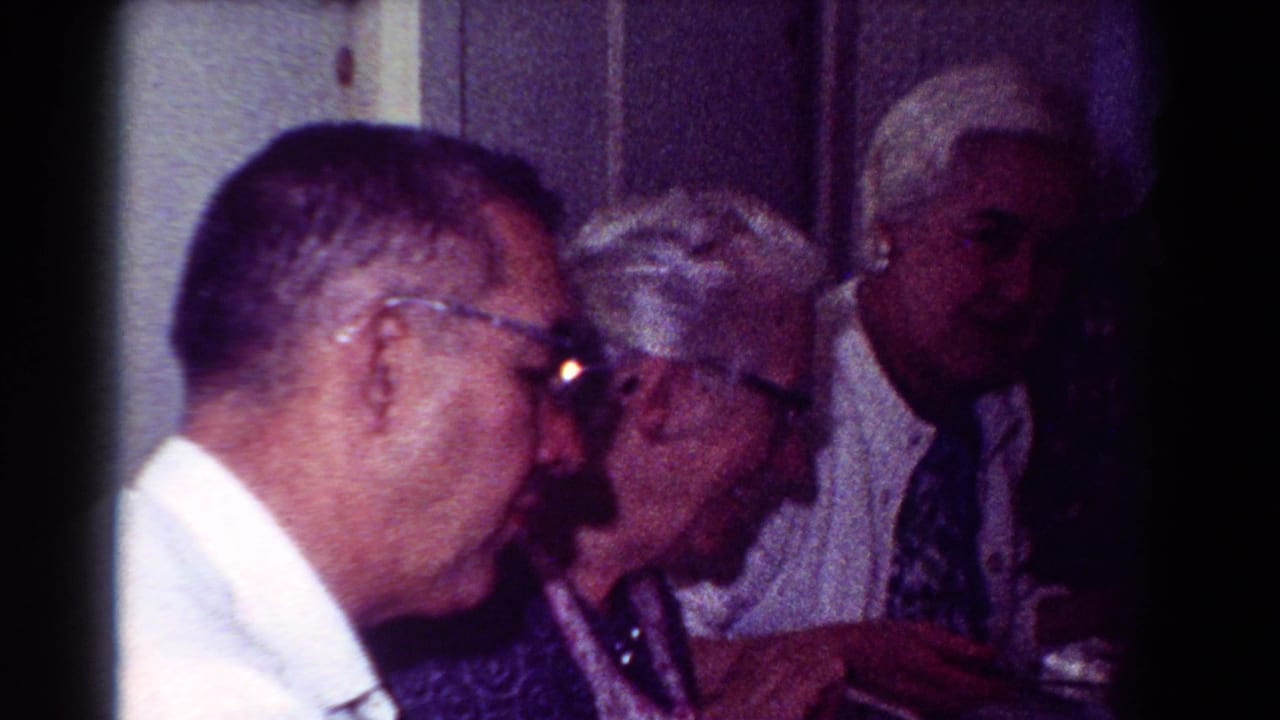1954 Toledo home-movie frame showing older adults in glasses gathered at a table during conversation.