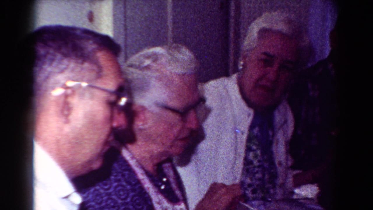 1954 Toledo home-movie frame with an elderly woman and older men seated close together in a private interior.