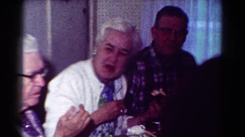1954 Toledo home-movie frame with a white-haired man gesturing near an older man in plaid inside a private room.