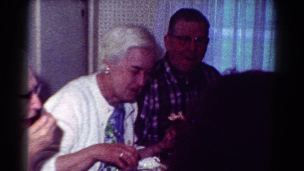 1954 Toledo home-movie frame showing adults at an indoor dining table, with a white-haired woman, a man in glasses, curtains, and patterned wallpaper.