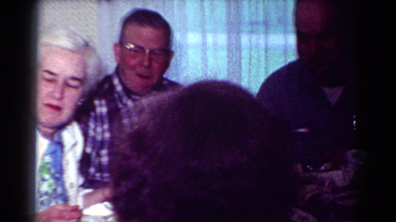 Adults gathered at a table with a white-haired woman, a man in plaid, and another person partly in shadow.