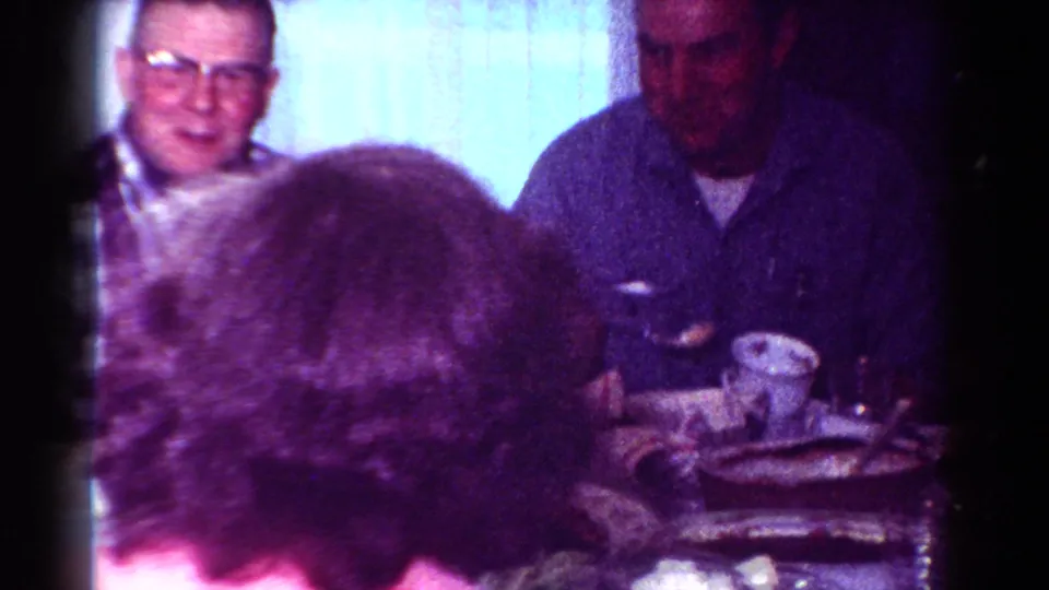 1954 Toledo home-movie frame showing a man in glasses at a crowded dining table with cups, plates, and other adults partly blocking the view.