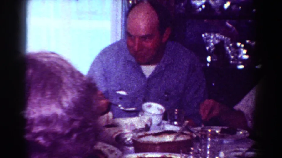 1954 Toledo indoor home-movie frame with a man in a blue shirt at a table with plates, glasses, cups, and shiny holiday-style decoration nearby.