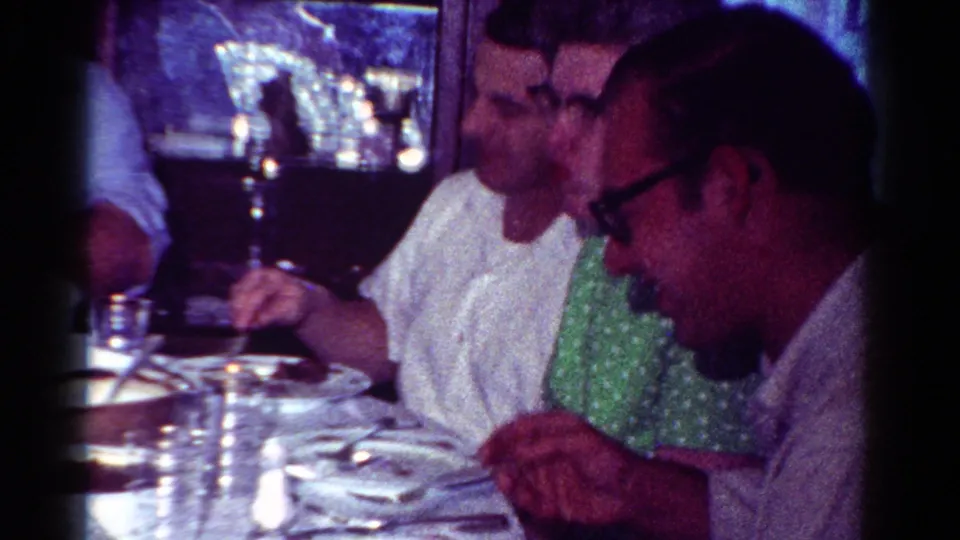 1954 Toledo home-movie frame showing adults seated in profile during an indoor family meal.