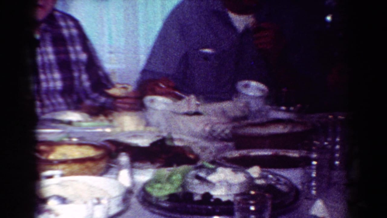 1954 Toledo home-movie frame of adults gathered at a crowded dining table with dishes, cups, and lace curtains in the background.