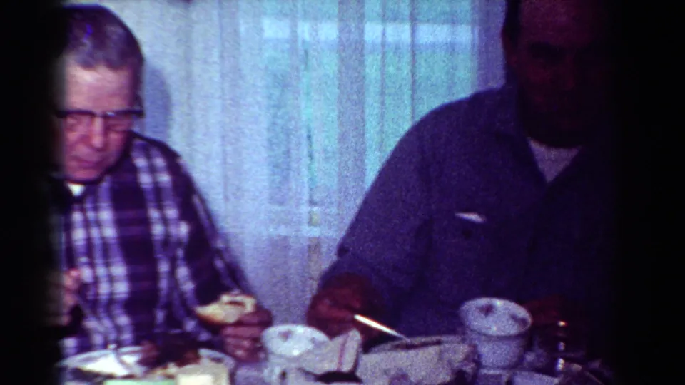 A man with glasses and a plaid shirt sits at the dining table with food, cups, napkins, and lace curtains visible in the home interior.