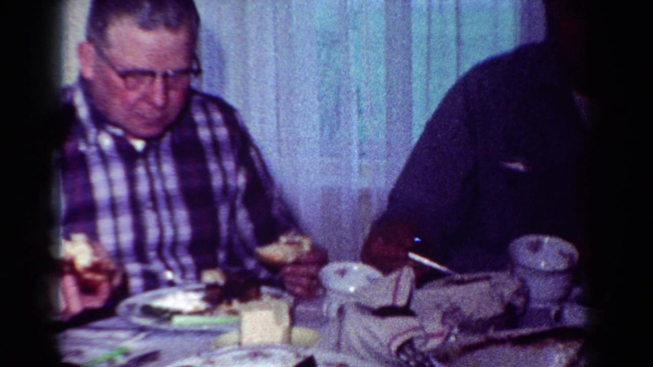 1954 Toledo home-movie frame of an older man in a plaid shirt and glasses seated at a dinner table with plates and cups.