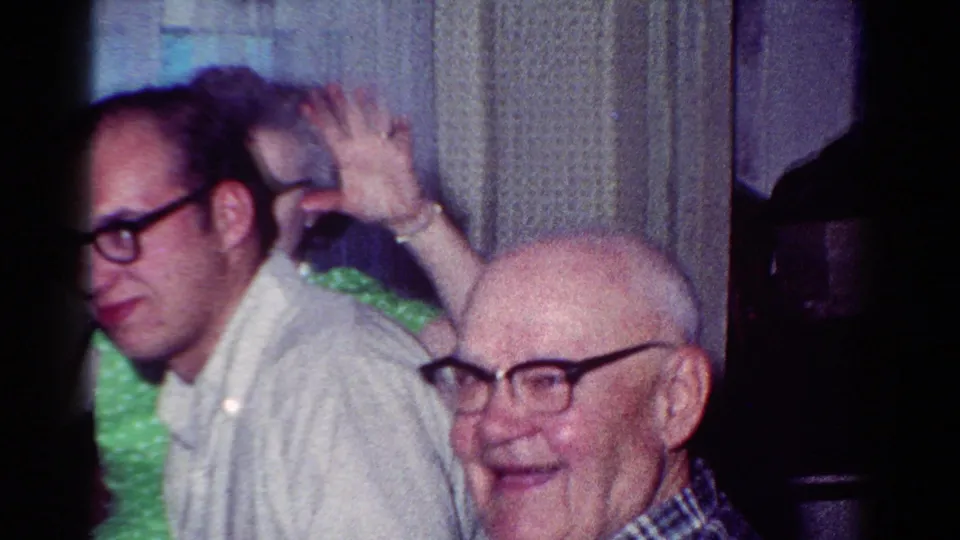 Two adult men in glasses sit close together at a 1954 Toledo family dinner, with one older man smiling toward the camera.