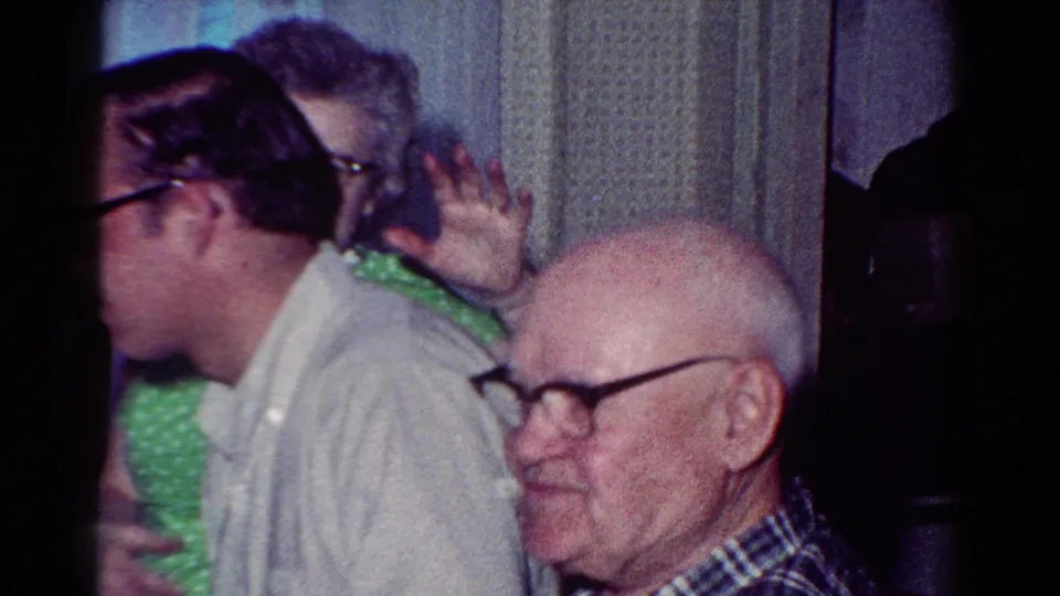 At a 1954 Toledo dinner, a man in glasses makes a playful face while an older man in a plaid shirt smiles nearby.