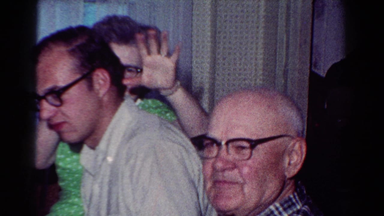 Older man in plaid sits beside a younger man while a woman in green waves behind them.
