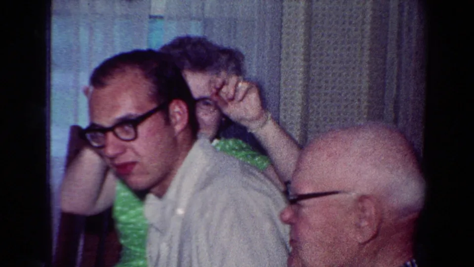 1954 Toledo indoor family scene with a younger man in glasses turning toward camera, a woman waving, and an older man in profile.