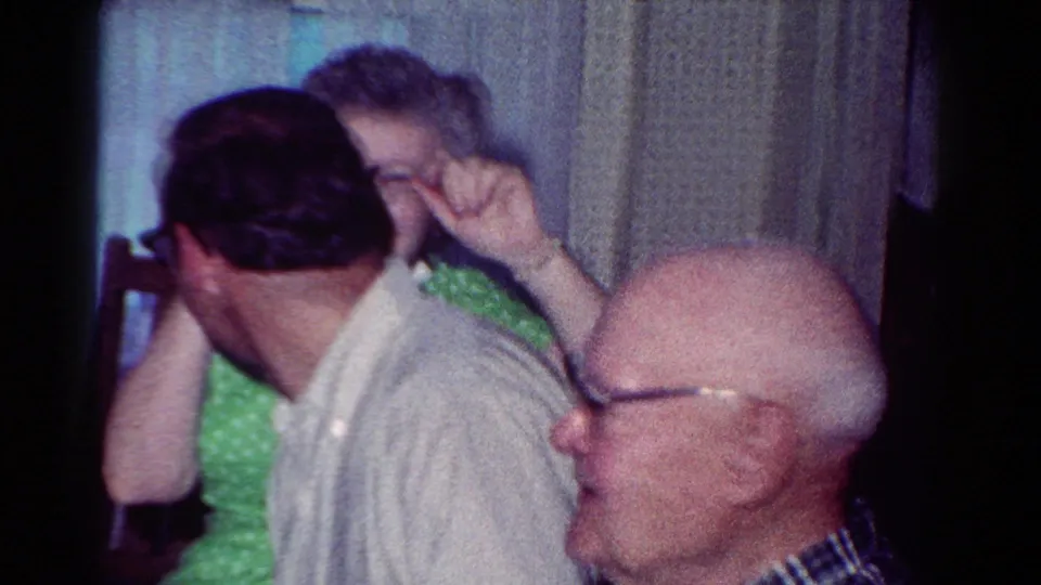 1954 Toledo home-movie frame with a younger man turned away, a woman in green behind him, and an older man in glasses at right.