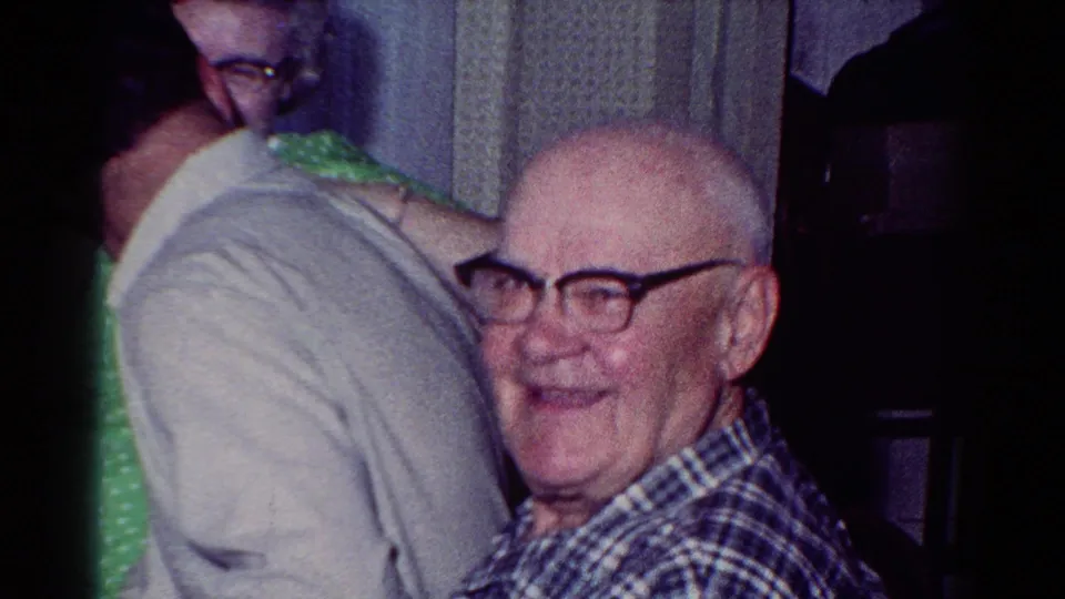 1954 Toledo family home-movie frame showing an older man in plaid smiling at the camera in a dim private interior.