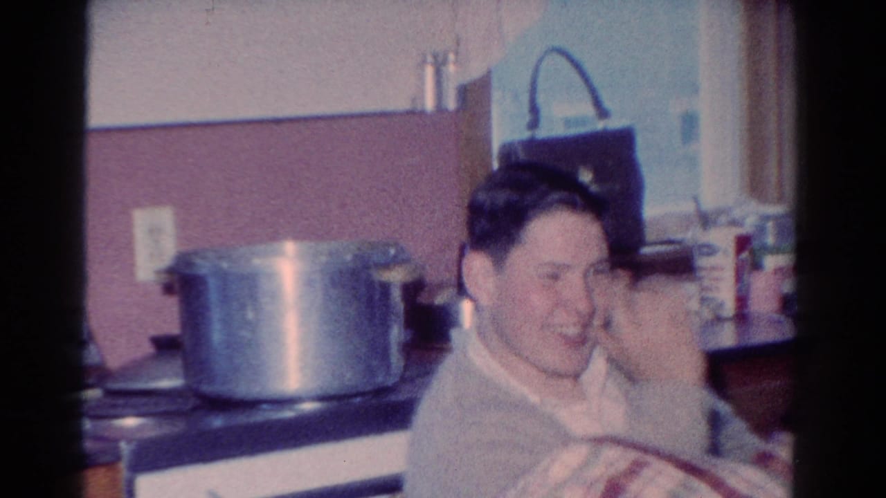 Man in a light sweater laughs in a kitchen beside a stove and large pot.