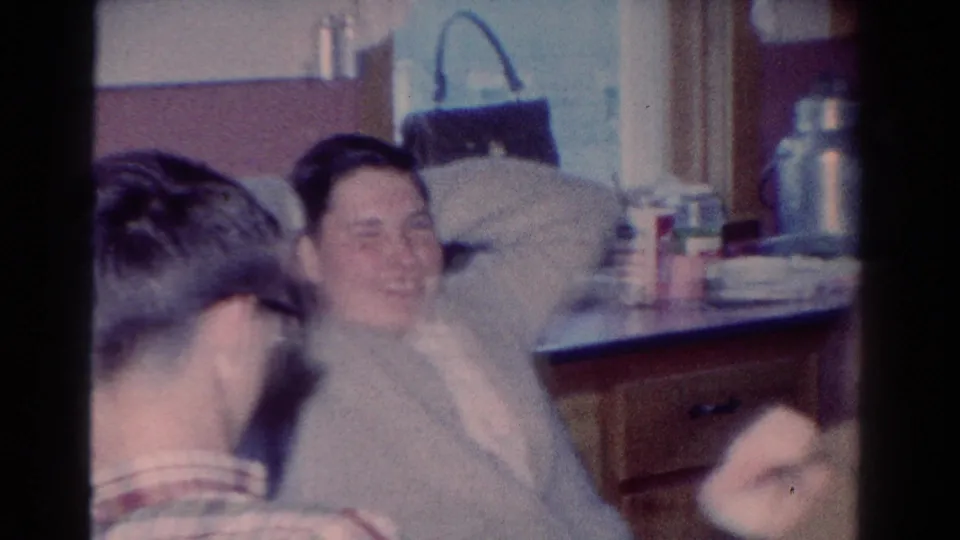 A 1954 Toledo home-movie frame shows a laughing man leaning back in a kitchen while a plaid-shirted man turns nearby.