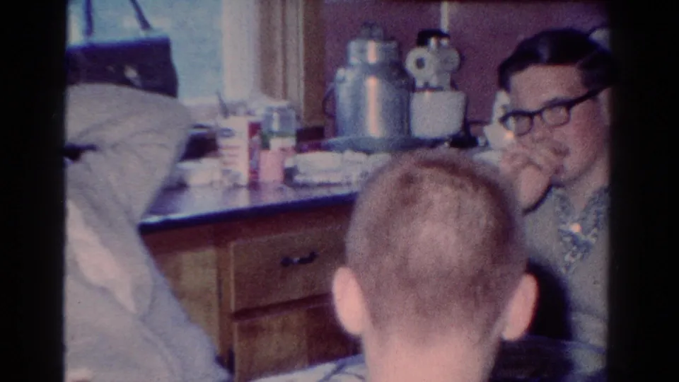 In a 1954 Toledo kitchen scene, a man with dark glasses smiles while others sit close around a counter with kettle and dishes.