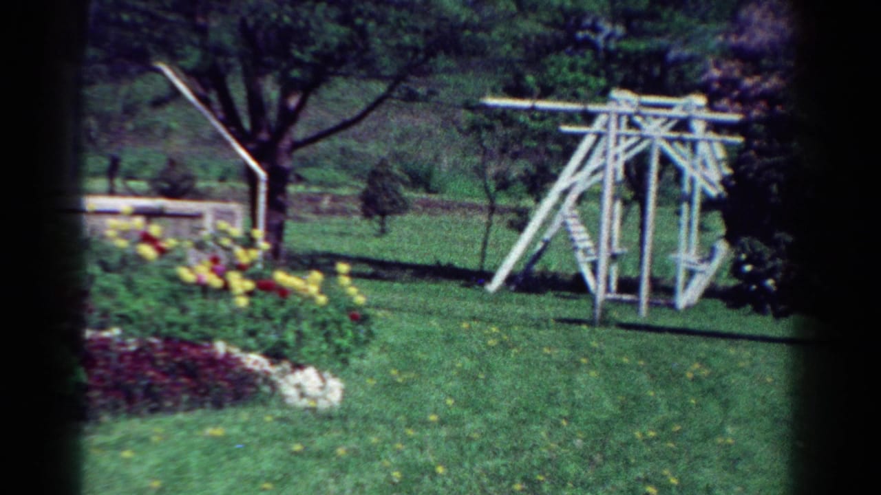 1954 Toledo backyard with a white wooden swing set, green lawn, flower bed, trees, and a low stone wall.