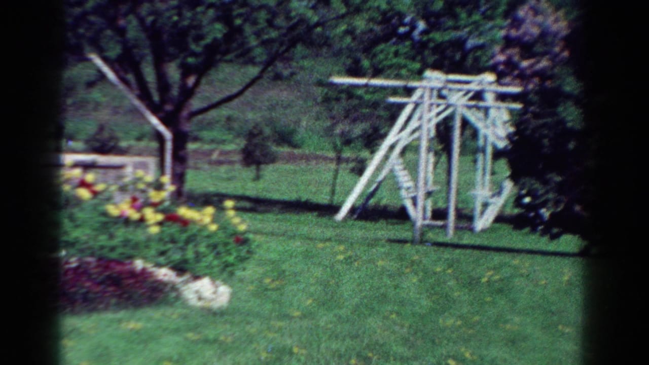 1954 Toledo home-movie frame showing a white backyard play structure across a green lawn with flowers and trees.