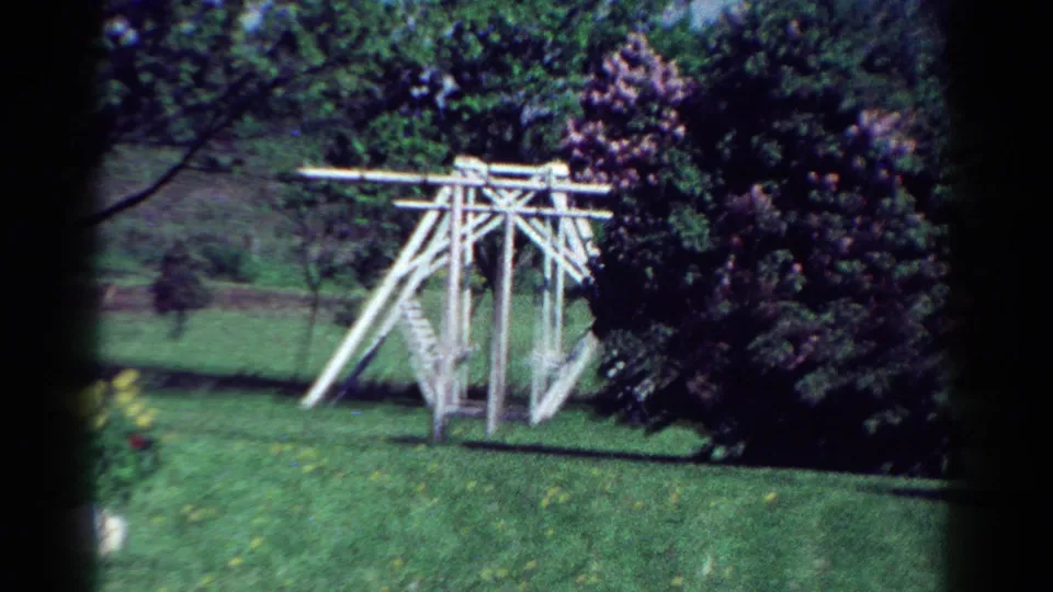 1954 Toledo backyard view with a white wooden swing set or play frame, mowed grass, trees, and dense shrubs.